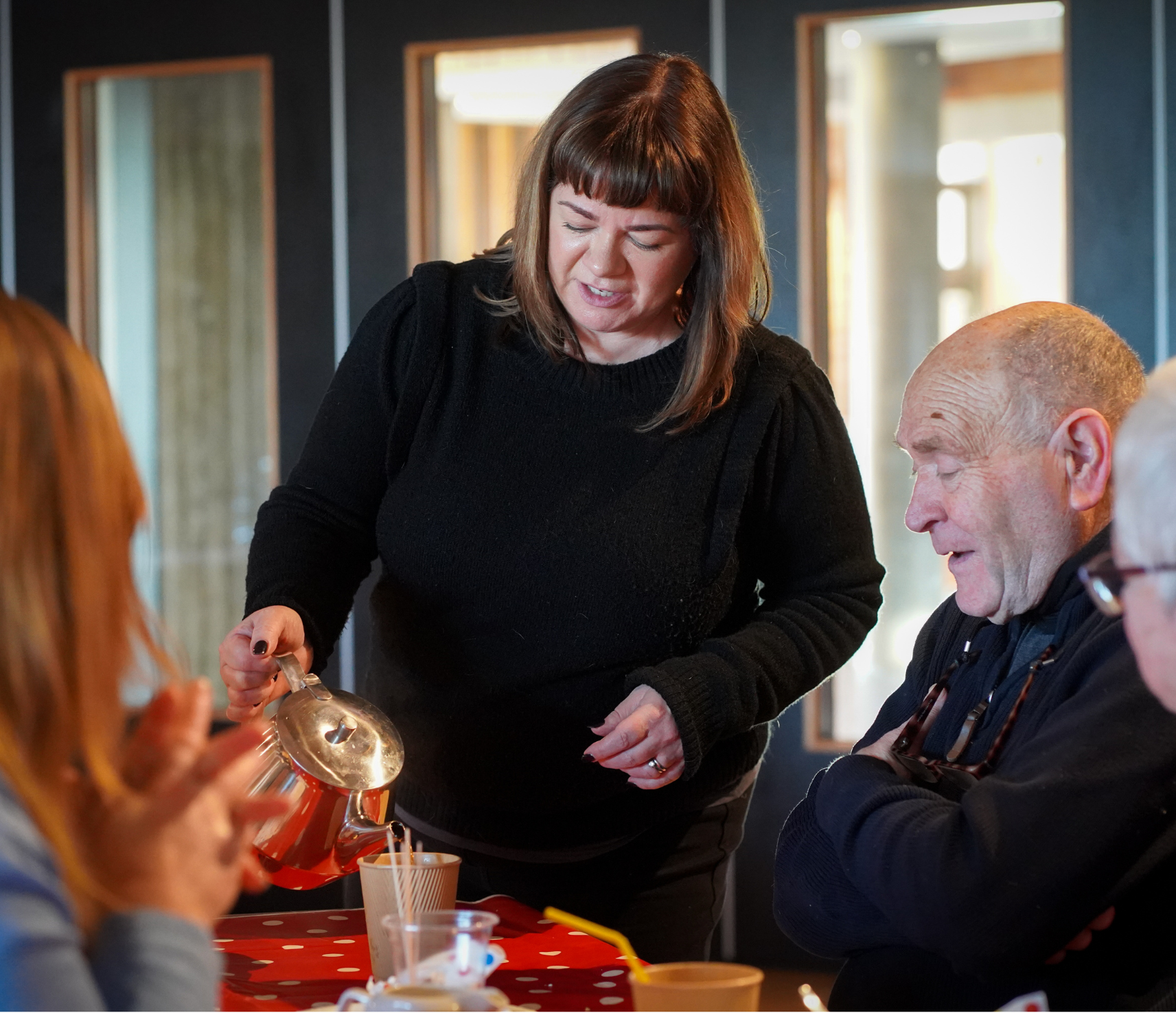 3 people sitting at a table while a woman in a black jumper pours tea from a teapot
