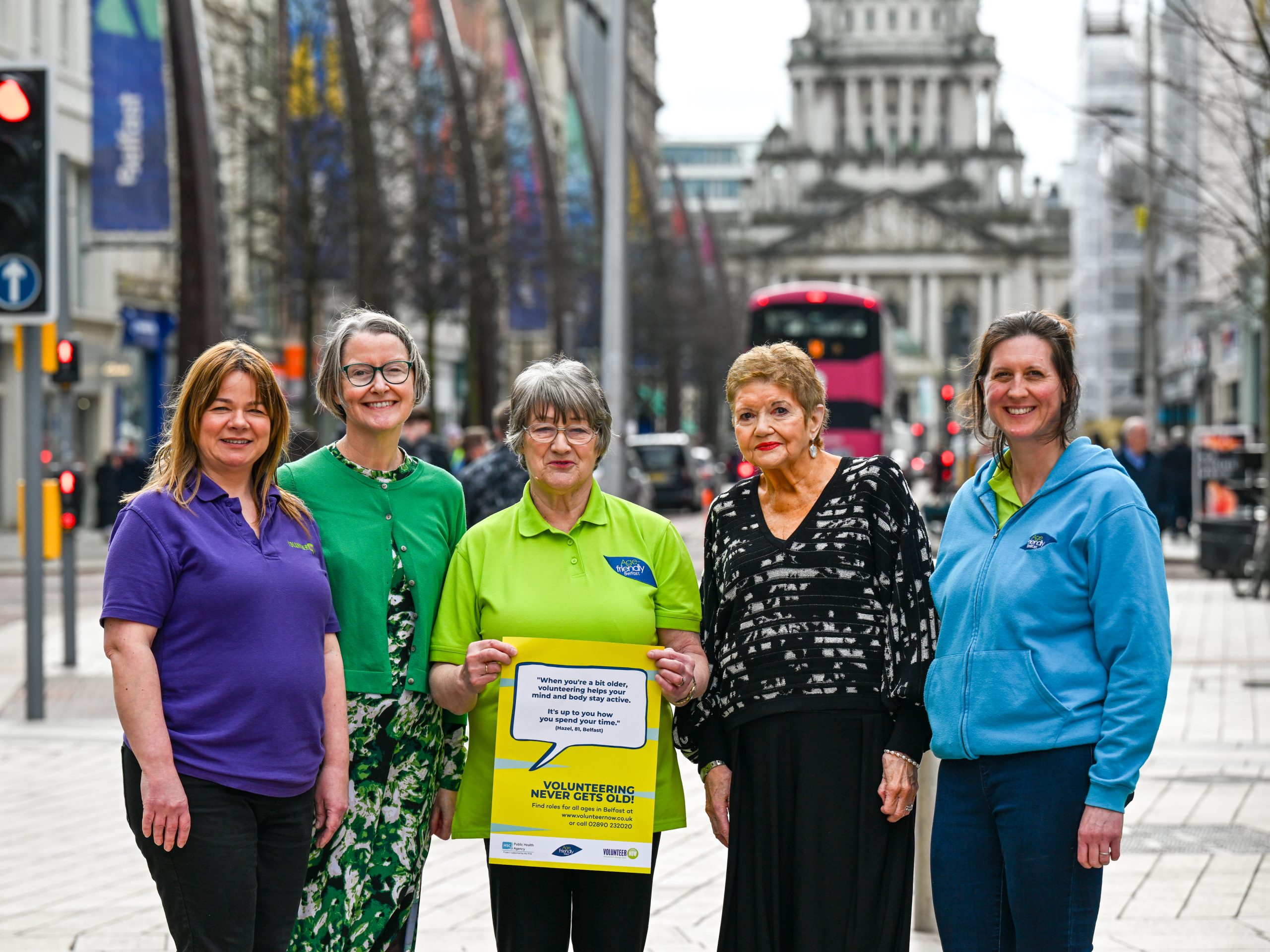 Photograph of 5 women wearing brightly coloured clothing in front of Belfast City Hall. One of the women is holding a poster advertising the Volunteering Never Gets Old campaign from Volunteer Now, Age Friendly Belfast and the Public Health Agency.